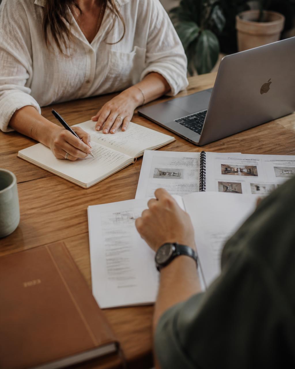 Two people working across a wooden table with notebooks, process documents, and a laptop, faces out of frame