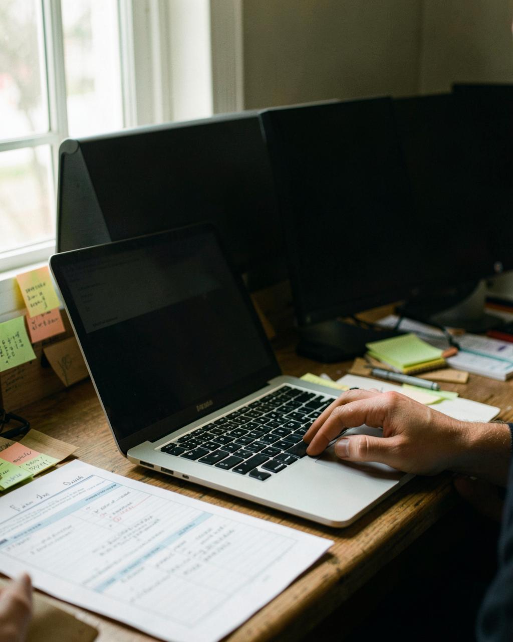 A laptop on a back-office desk covered in sticky notes, printed spreadsheets and a half-finished checklist